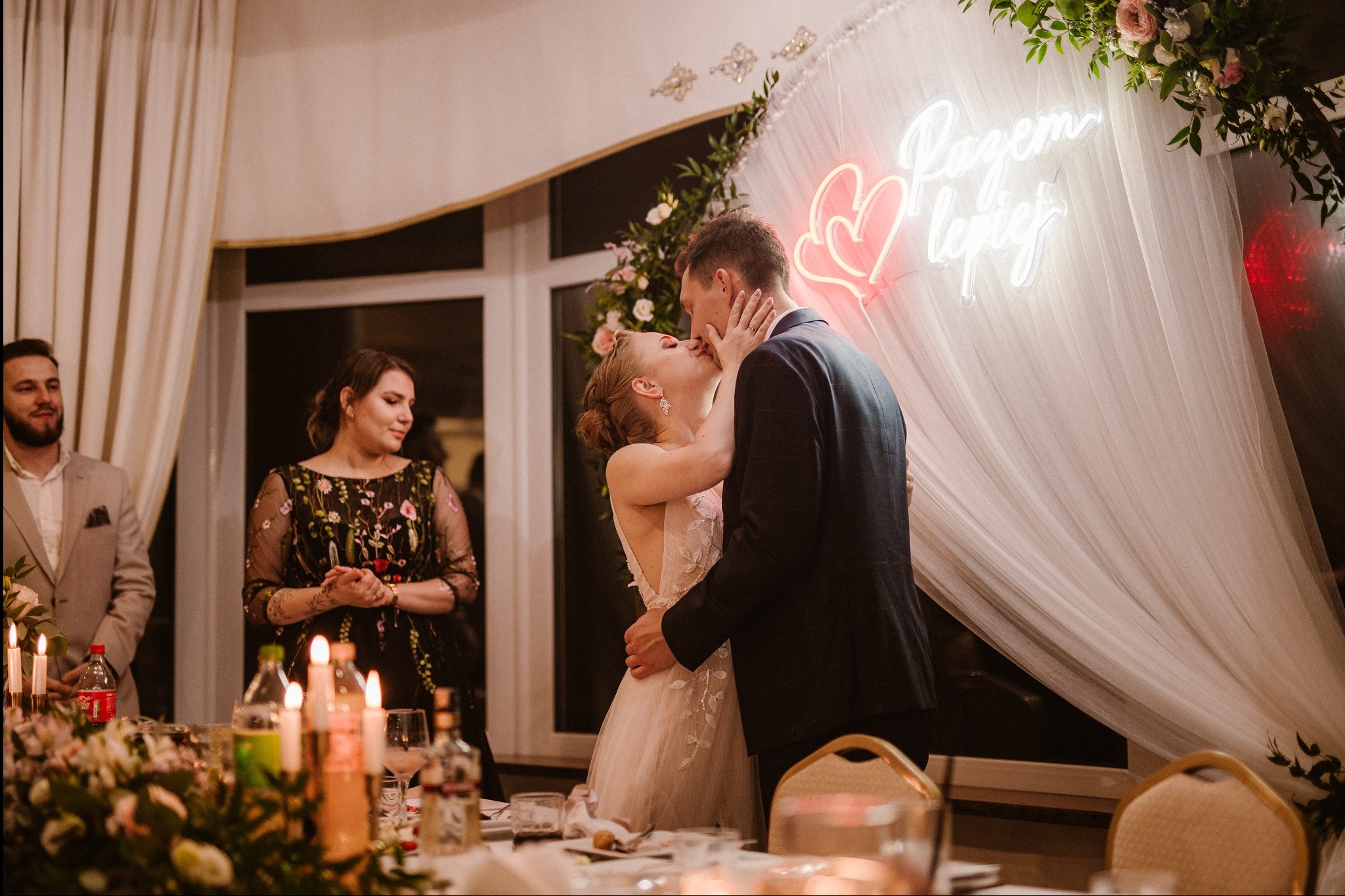 Pocałunek pary młodej na tle dekoracji ze światłem neonowym i kwiatami. Na stole dekoracje, kwiaty i butelki. W tle goście weselni.