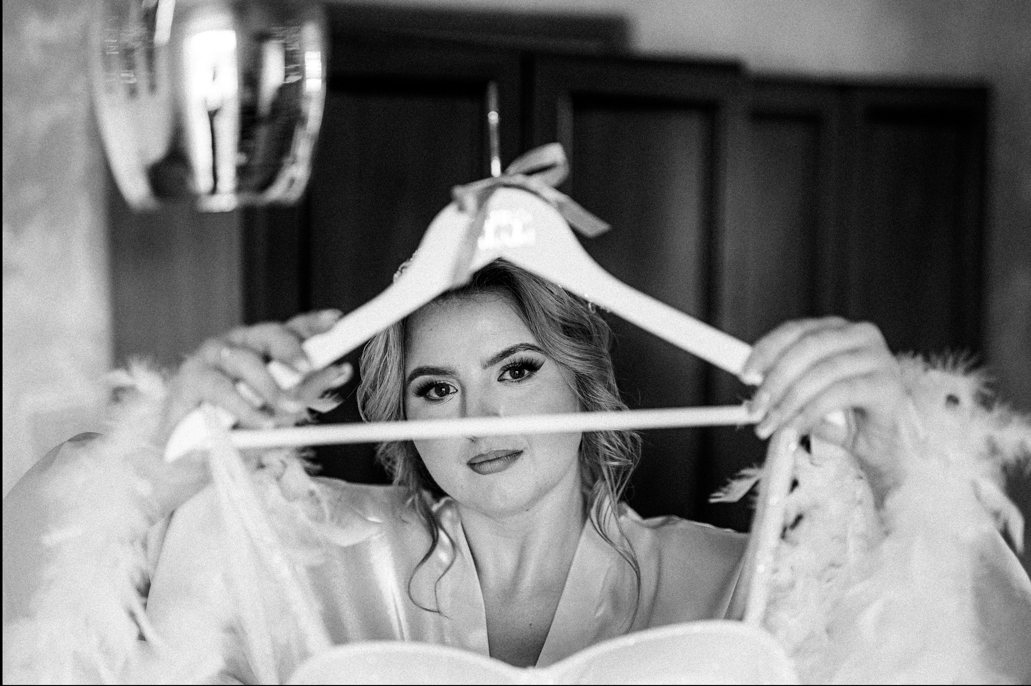 Czarno-białe ujęcie panny młodej trzymającej na wieszaku suknię ślubną z piórami, spoglądającej w obiektyw, z rozproszonym światłem w tle.