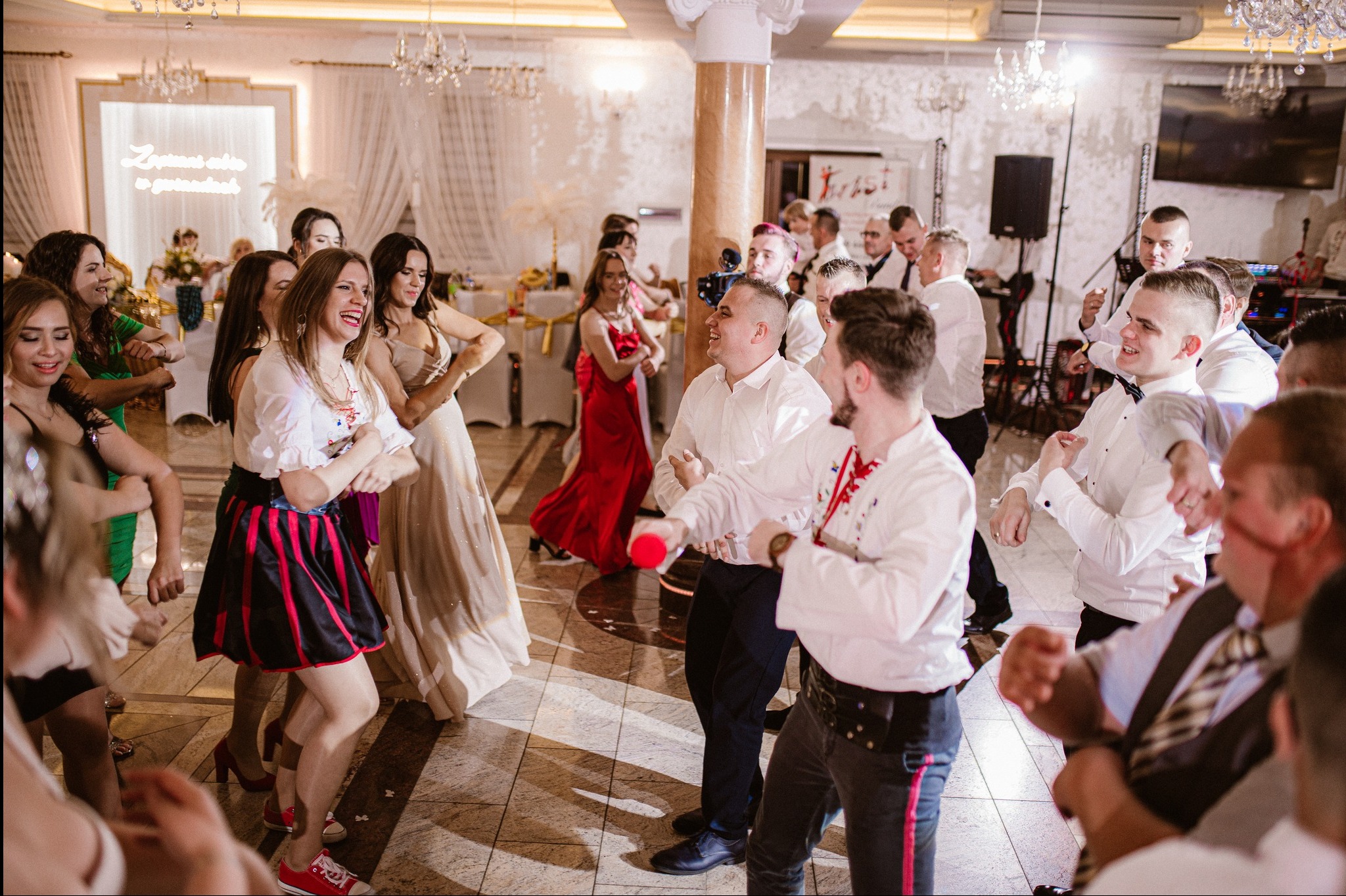 Radosna zabawa weselna w sali bankietowej: tańczący goście w różnorodnych strojach, oświetlona scena i kryształowe żyrandole tworzą świąteczny klimat.
