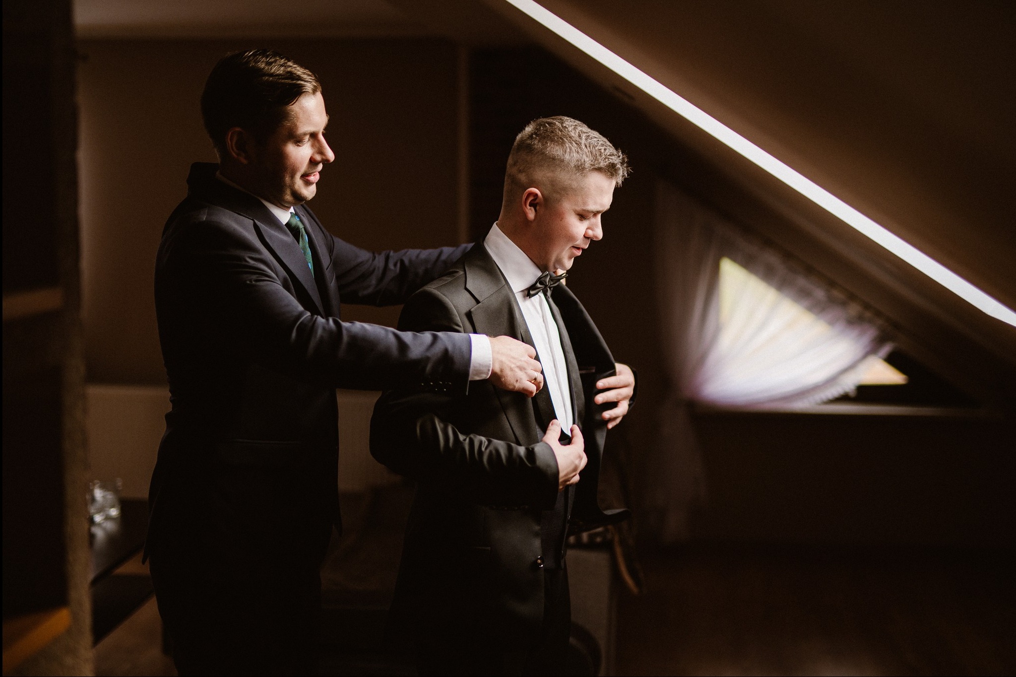 Mężczyzna w garniturze pomaga innemu mężczyźnie dopiąć marynarkę przed ślubem, w tle okno z zasłonami i ukośny sufit z oświetleniem.