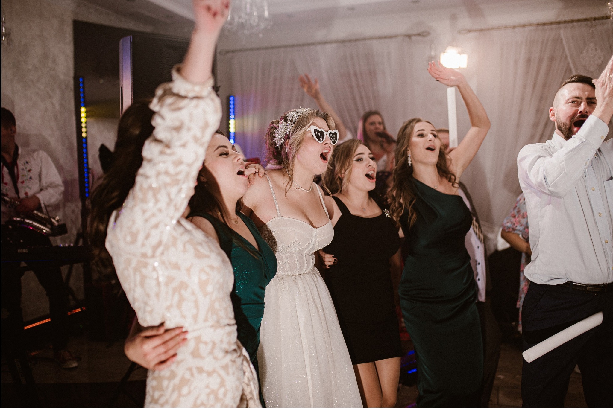 Radosna panna młoda w okularach w kształcie serc i goście weselni podczas zabawy, w tle muzycy i dekoracje świetlne.