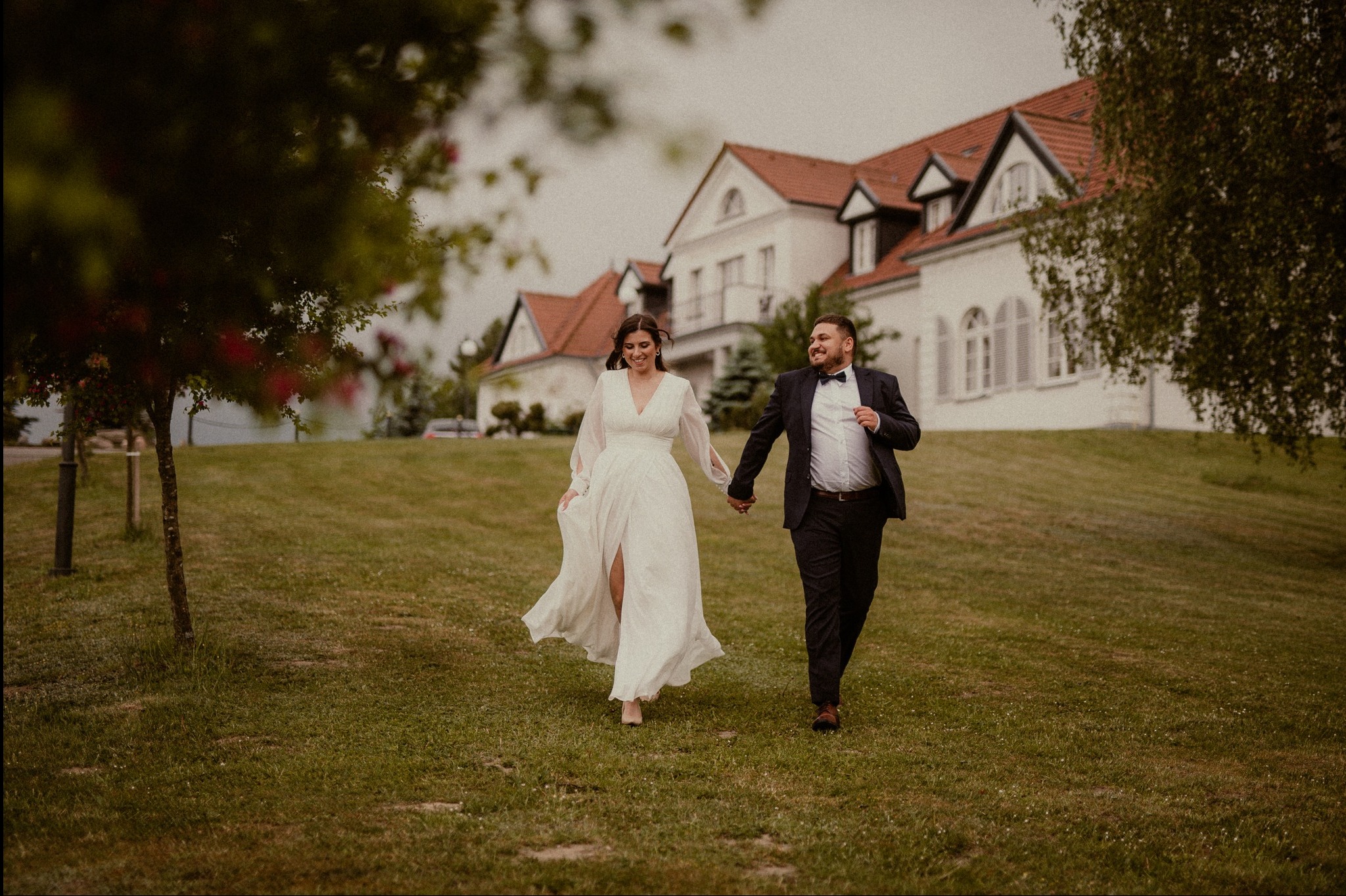 Uśmiechnięta para młoda, w ruchu, trzymająca się za ręce na tle białego budynku z czerwonym dachem, w plenerze. Ona w zwiewnej sukni, on w garniturze.