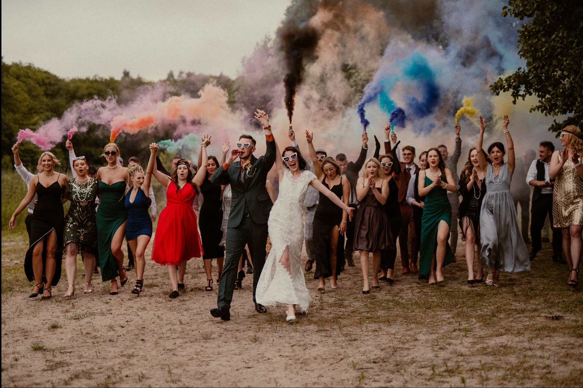 Para młoda i goście weselni z kolorowymi dymami na tle zieleni, wesoła atmosfera, nietypowe okulary w kształcie serc, plenerowa sesja zdjęciowa.