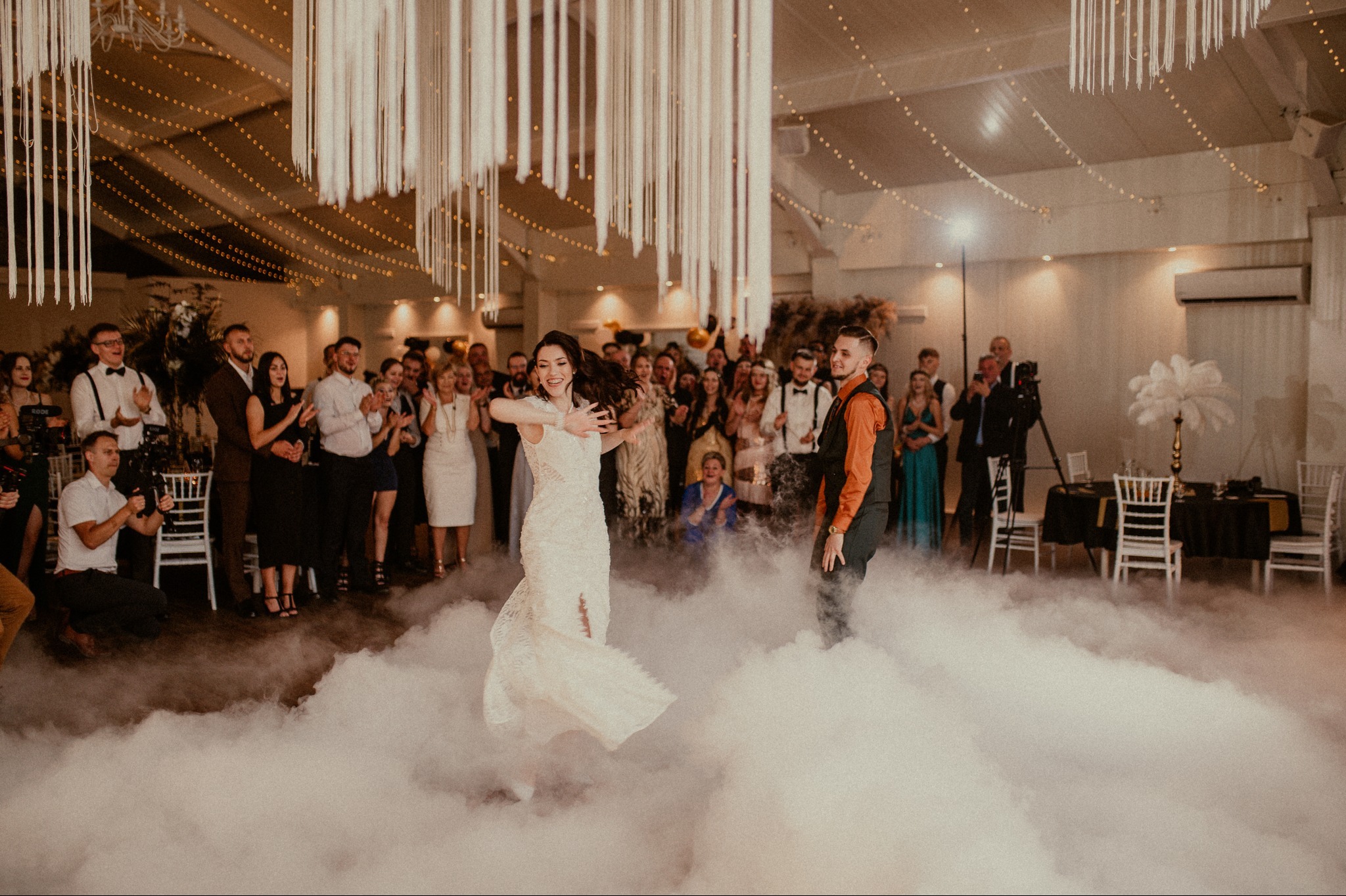 Pierwszy taniec pary młodej w dymie na tle gości weselnych. Panna młoda w koronkowej sukni, pan młody w kamizelce. Dekoracje sali z wiszącymi elementami.
