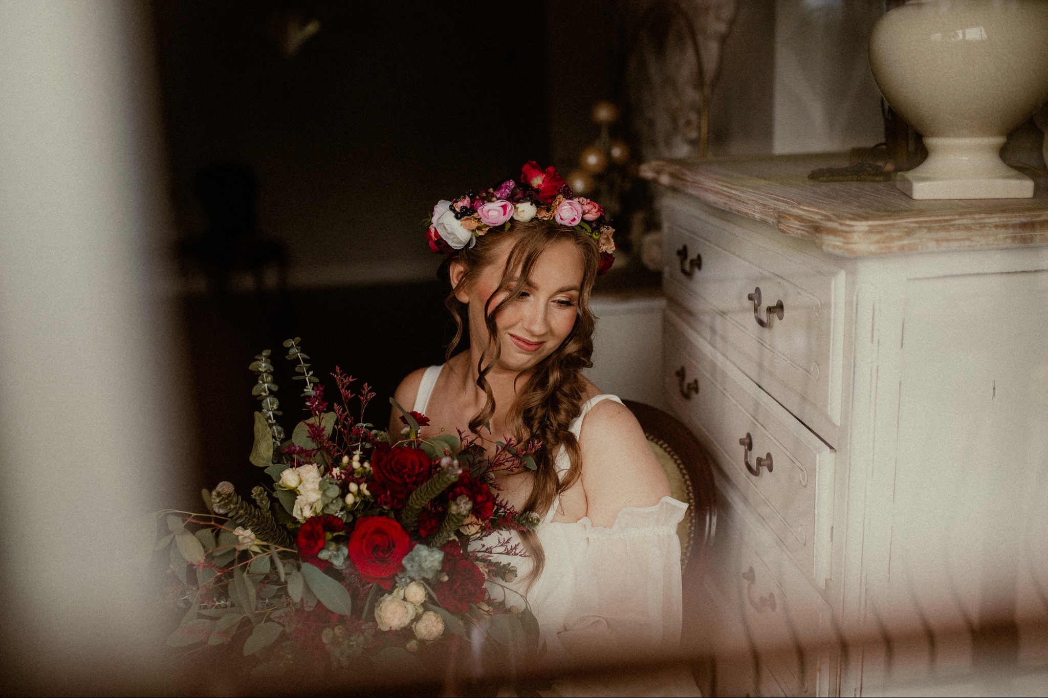 Uśmiechnięta kobieta z wiankiem i bukietem w rękach, delikatne światło, w tle biała komoda. Romantyczna stylizacja i naturalny makijaż.