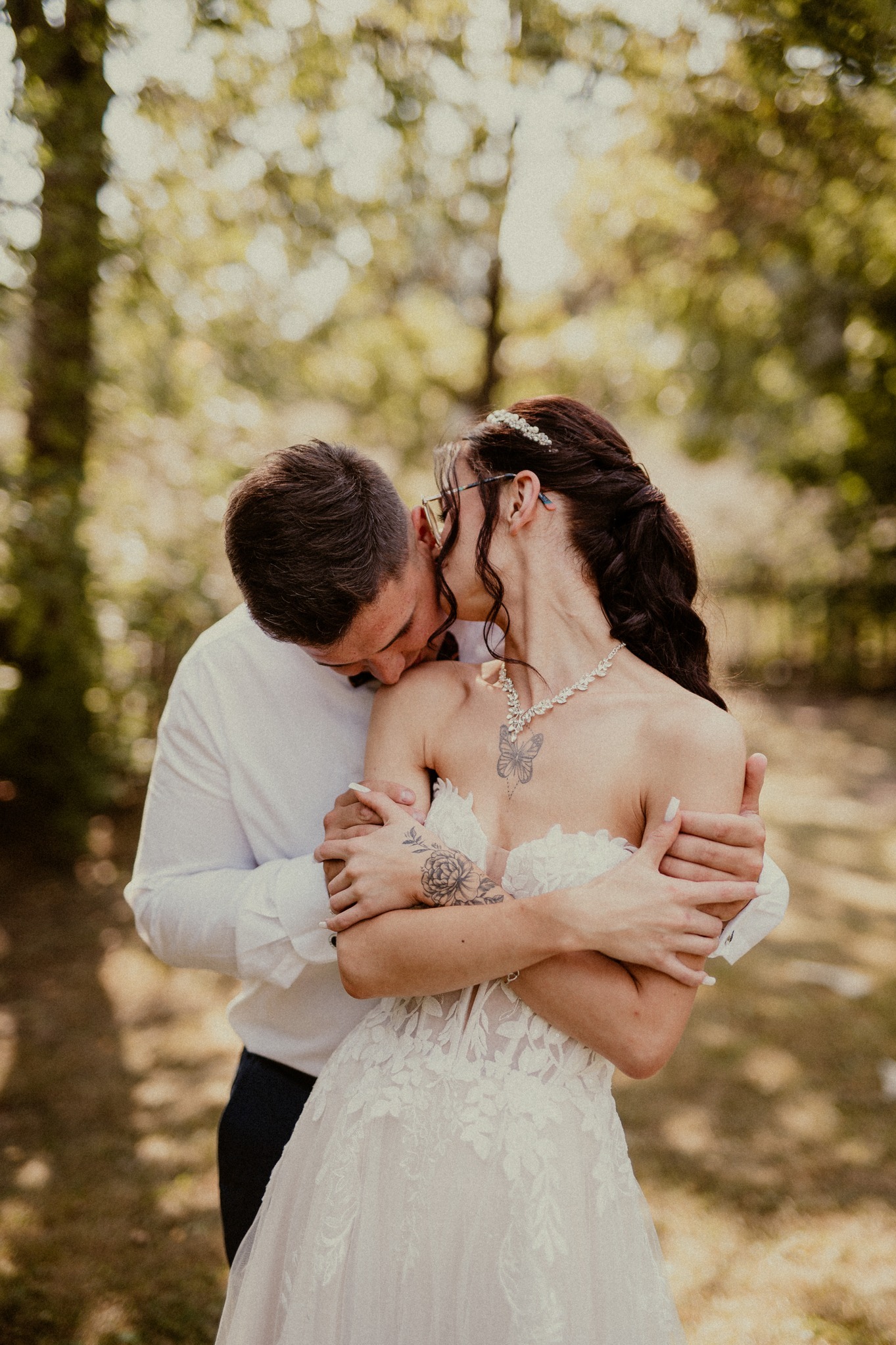 Para młoda w objęciach na tle zieleni. Panna młoda w sukni z koronką, z tatuażami na ramionach i naszyjnikiem. Pan młody w białej koszuli i muszce całuje szyję kobiety.