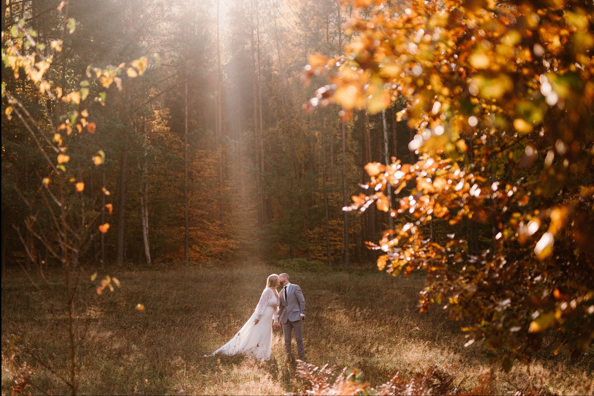Para młoda w jesiennym lesie, promienie słońca przeswitują przez drzewa. Panna młoda w koronkowej sukni, pan młody w szarym garniturze. Romantyczna sceneria.