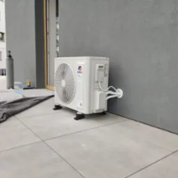 Zewnętrzna jednostka klimatyzacji Gree, zamontowana na szarym balkonie, widoczna butla z gazem i zabezpieczenie podłoża. Instalacja na tle budynku.