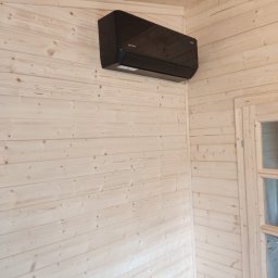 Paweł Maniecki AIRPRO - Czarna klimatyzacja ścienna w pomieszczeniu wykończonym jasnym drewnem, widoczna część okna z drewnianą ramą. Minimalistyczny design.