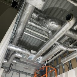 Paweł Maniecki AIRPRO - Srebrne, zaizolowane rury wentylacyjne pod sufitem, widoczne z podnośnika koszowego. Instalacja w trakcie montażu, sufit z blachy trapezowej.