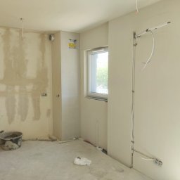 Paweł Maniecki AIRPRO - Wnętrze w trakcie remontu: ściany z instalacjami elektrycznymi i hydraulicznymi, widoczny kubeł budowlany, okno. Etap prac instalacyjnych przed wykończeniem.