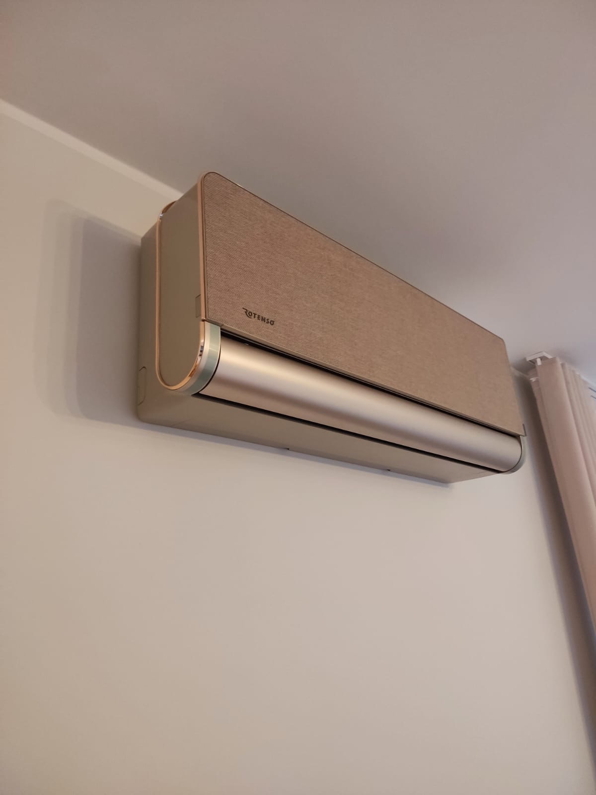 Zamontowana, beżowa klimatyzacja Rotenso z teksturowanym panelem frontowym i srebrnym paskiem, widoczna na białej ścianie w jasnym wnętrzu.