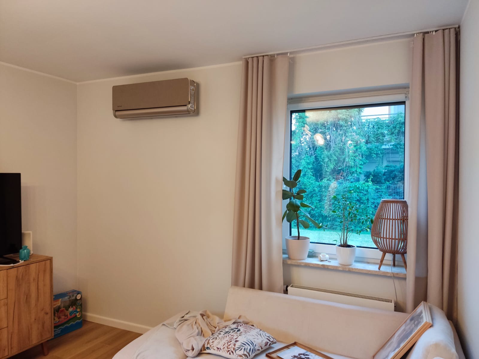 Klimatyzator ścienny w odcieniach beżu, zamontowany w nowoczesnym salonie z oknem i roślinami. Widoczna część sofy i komody z telewizorem.