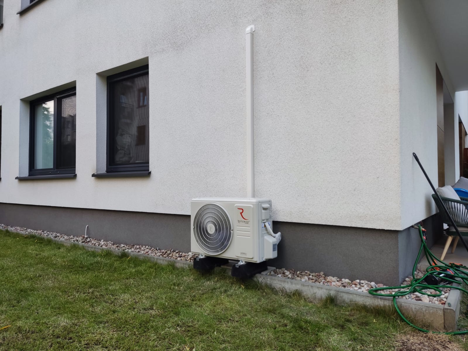 Biała jednostka zewnętrzna klimatyzacji Rotenso zamontowana na elewacji domu z trawnikiem. Widoczne okna i rura odprowadzająca. Ujęcie z bliska, z poziomu gruntu.