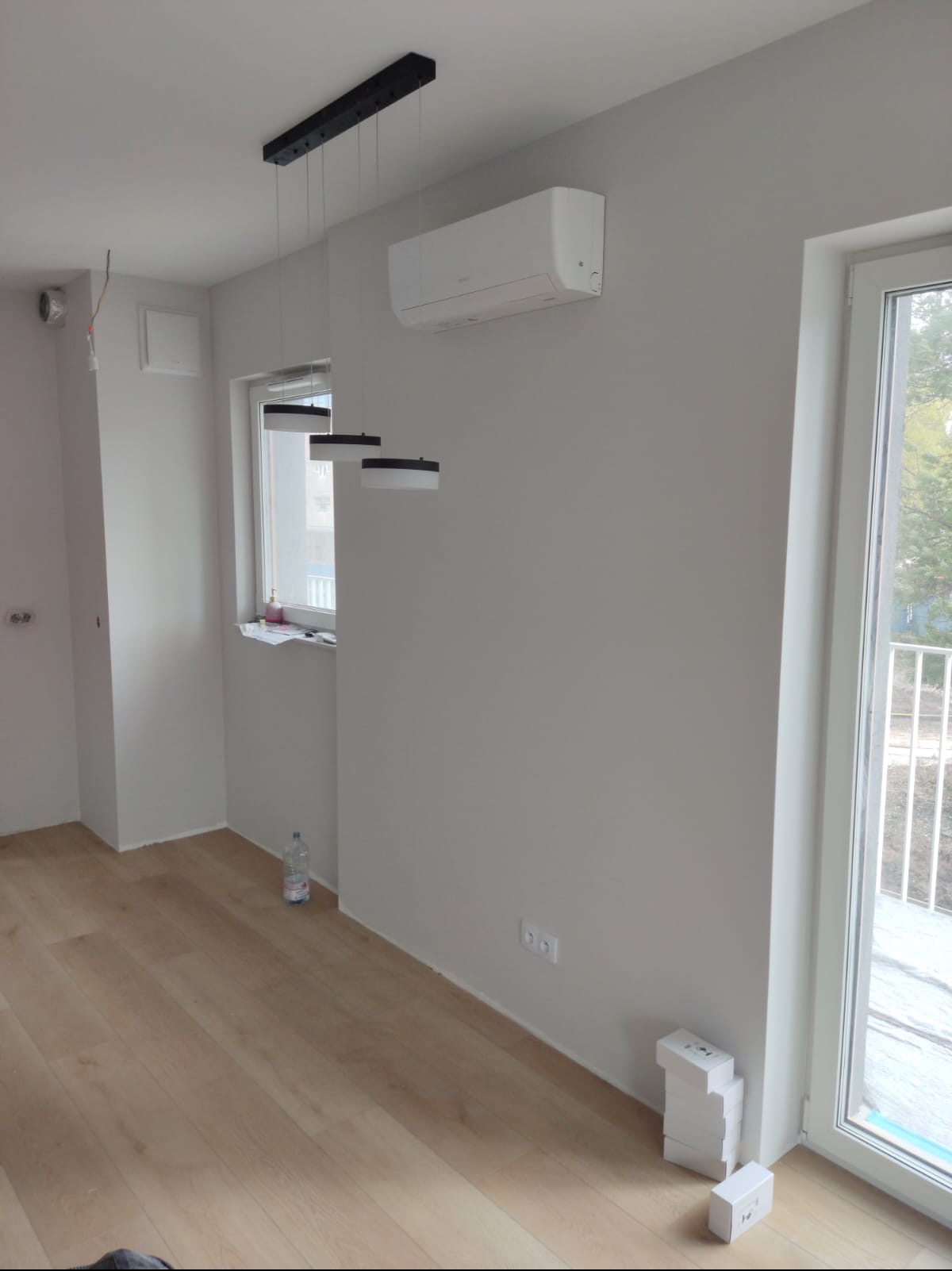 Wnętrze po montażu klimatyzacji ściennej. Widoczny biały klimatyzator, nowoczesna lampa wisząca i świeżo pomalowane ściany w odcieniach szarości. Podłoga panelowa.