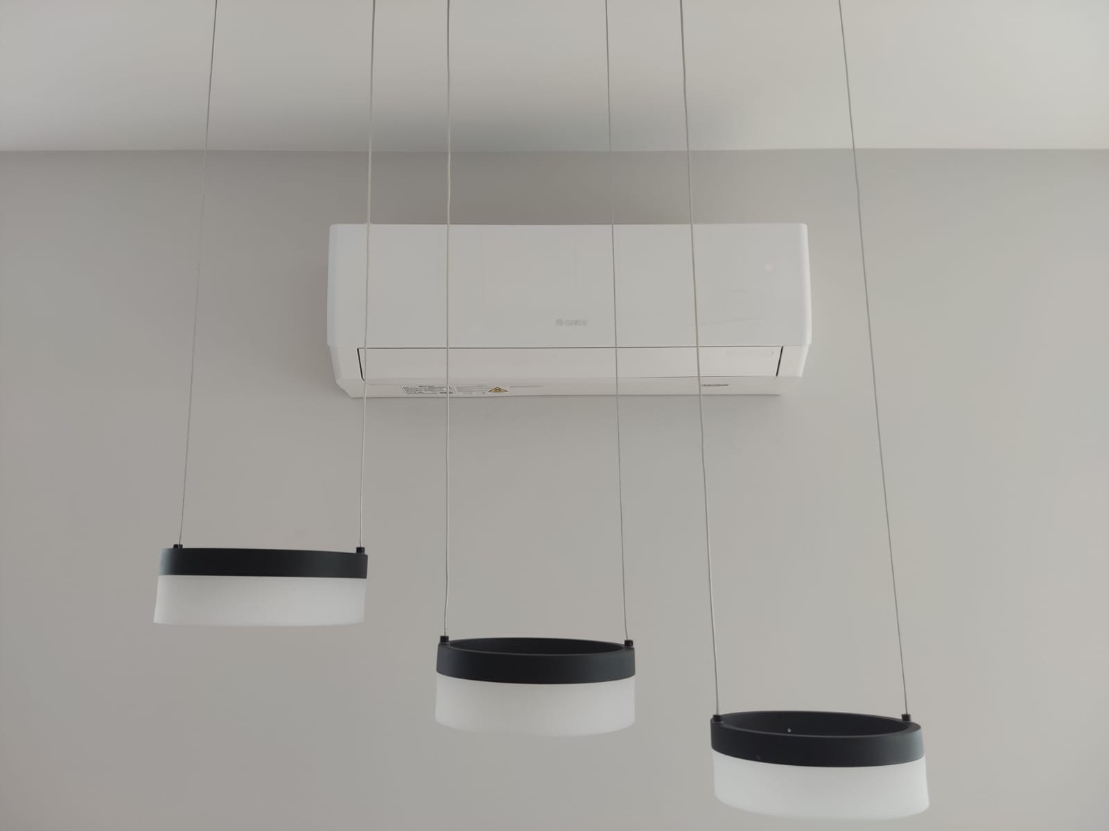 Biała klimatyzacja marki Gree zamontowana na ścianie, widoczna spod trzech nowoczesnych lamp wiszących z czarnymi i białymi elementami.