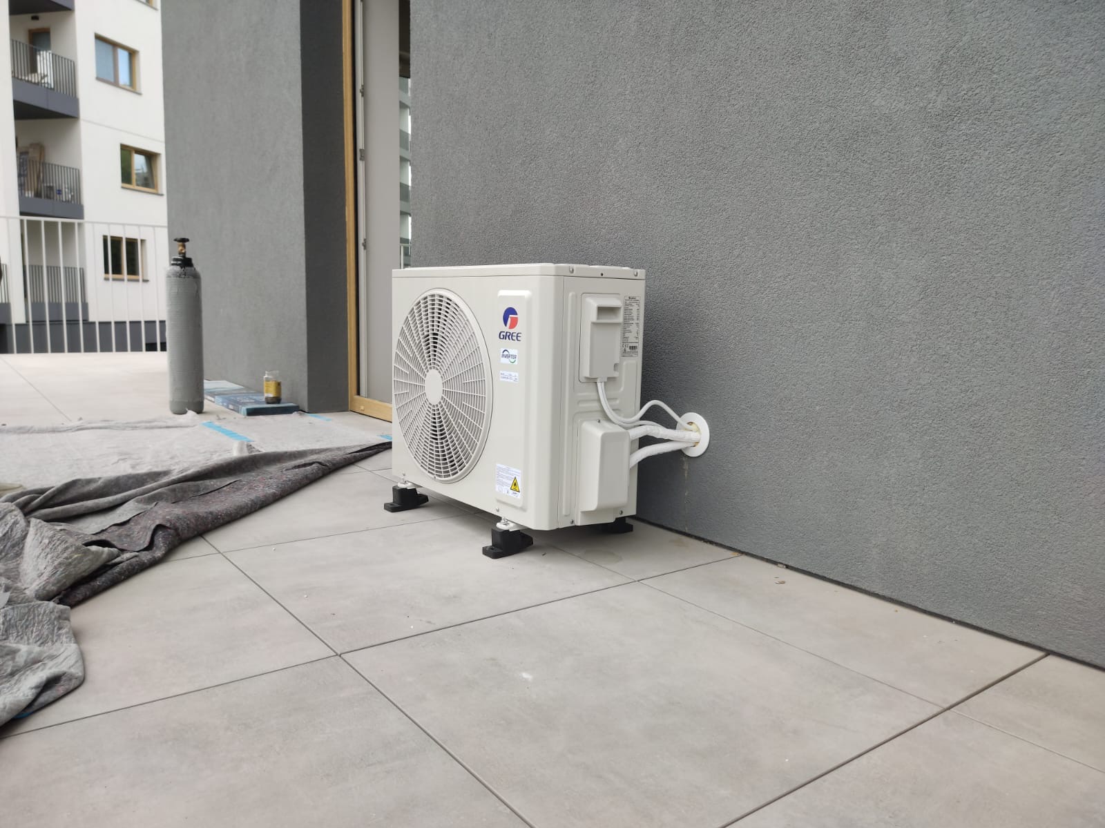 Zewnętrzna jednostka klimatyzacji Gree, zamontowana na szarym balkonie, widoczna butla z gazem i zabezpieczenie podłoża. Instalacja na tle budynku.
