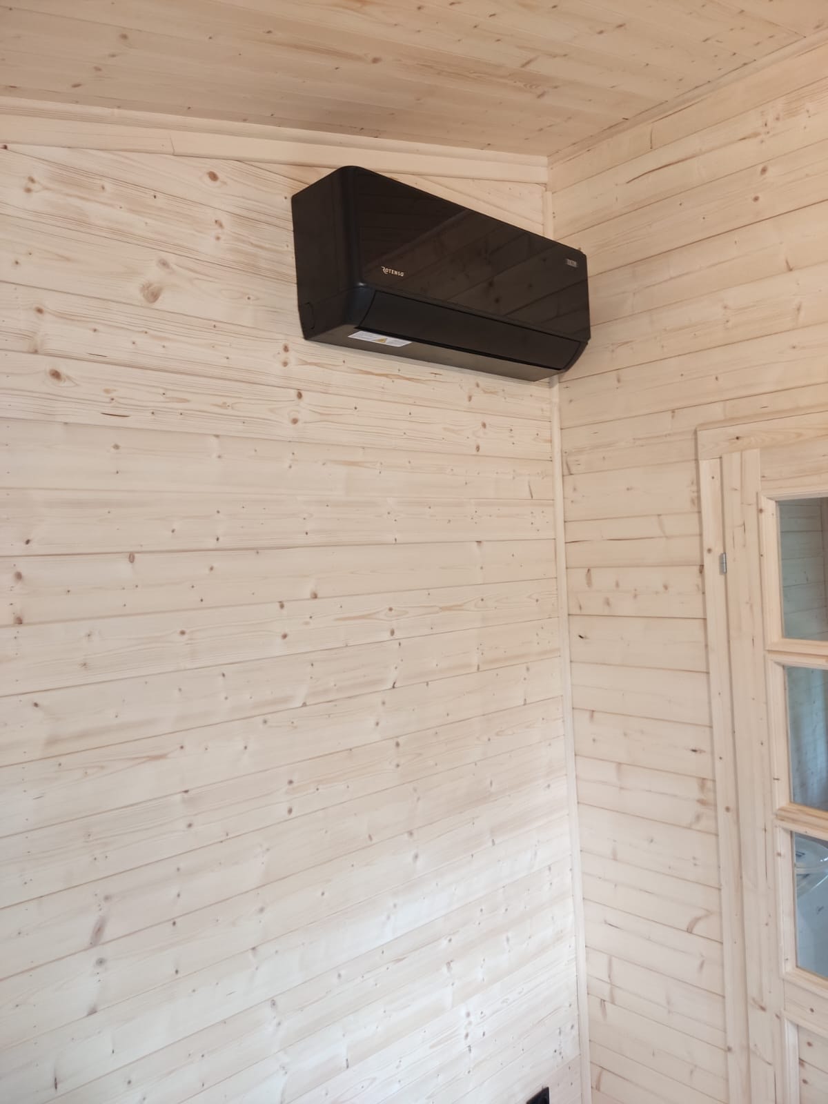 Czarna klimatyzacja ścienna w pomieszczeniu wykończonym jasnym drewnem, widoczna część okna z drewnianą ramą. Minimalistyczny design.