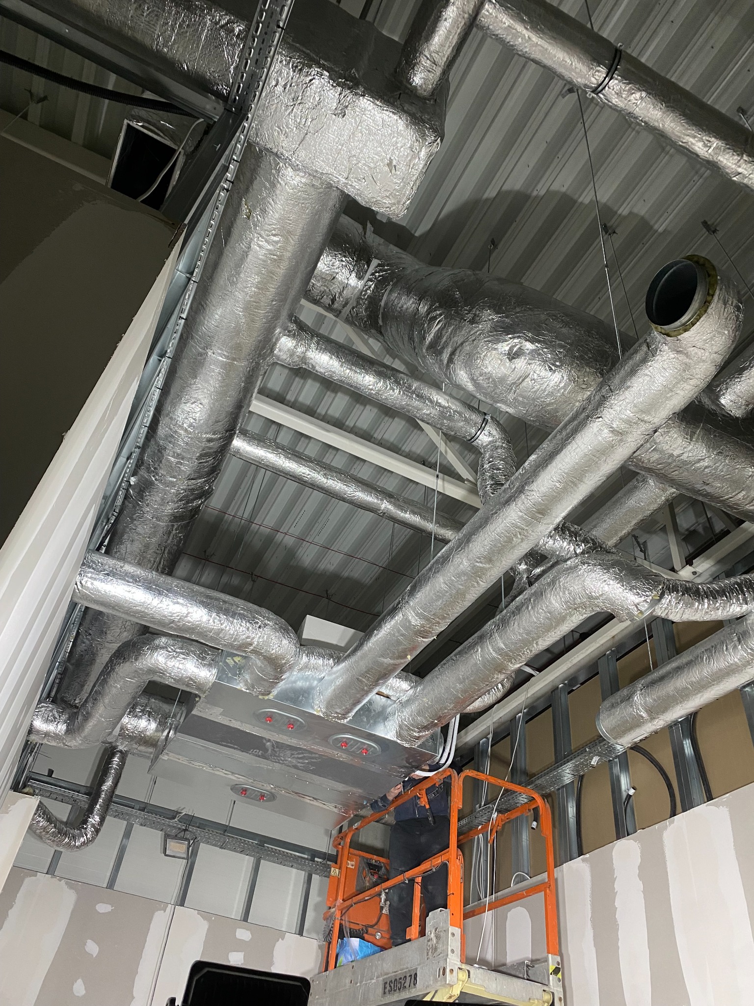 Srebrne, zaizolowane rury wentylacyjne pod sufitem, widoczne z podnośnika koszowego. Instalacja w trakcie montażu, sufit z blachy trapezowej.