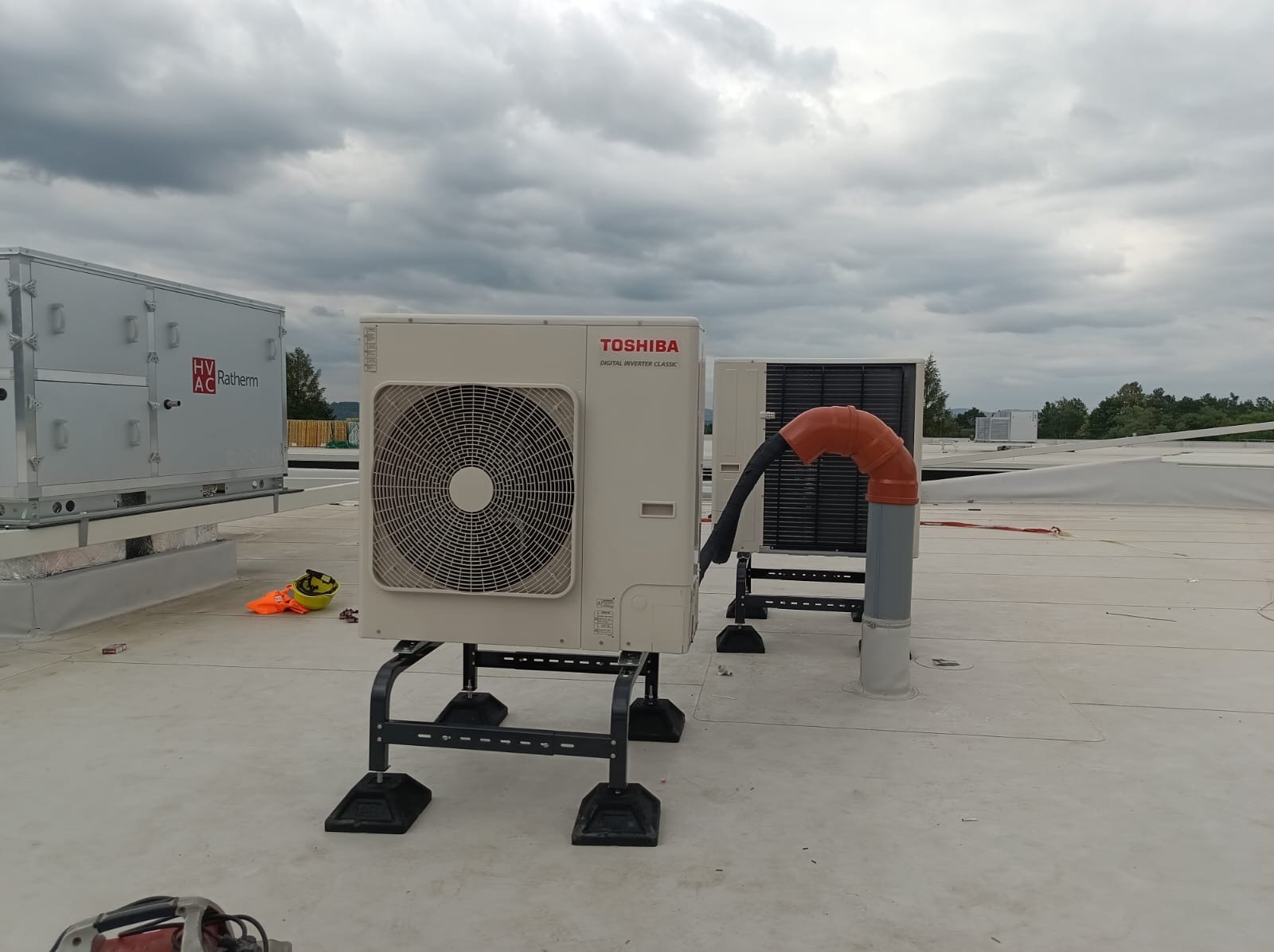 Biała jednostka zewnętrzna klimatyzacji Toshiba na dachu, z widocznymi elementami instalacji i pochmurnym niebem w tle. Widoczne narzędzia pracy.