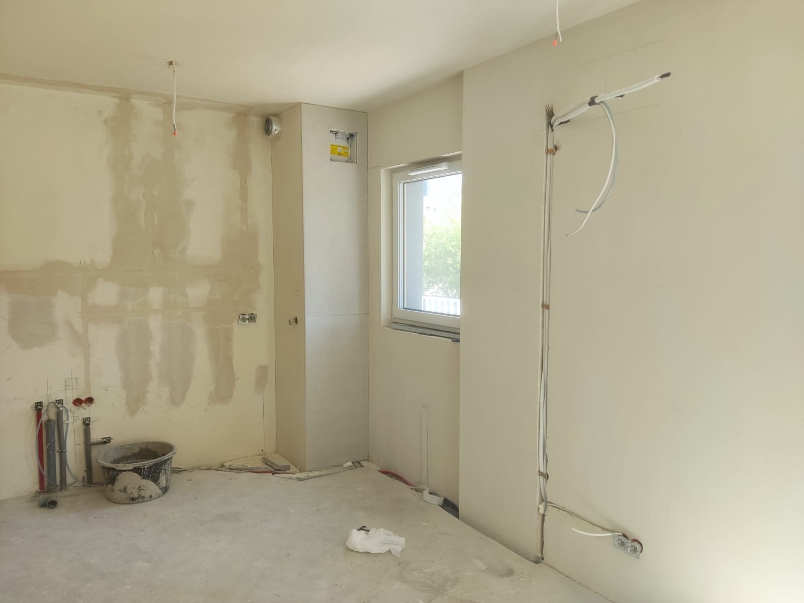 Wnętrze w trakcie remontu: ściany z instalacjami elektrycznymi i hydraulicznymi, widoczny kubeł budowlany, okno. Etap prac instalacyjnych przed wykończeniem.