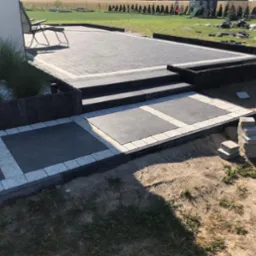 Nowoczesny taras z kostki brukowej w odcieniach szarości, z niskim murkiem i schodami. W tle widoczny trawnik i rząd drzew. Elementy dekoracyjne z białego kamienia.