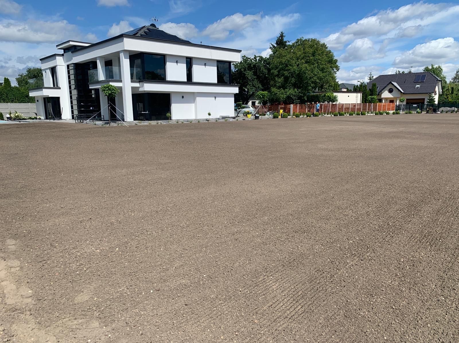 Przygotowany teren pod trawnik, widoczna świeżo wyrównana ziemia przed nowoczesnym domem z ogrodem. Ślady po bronowaniu.