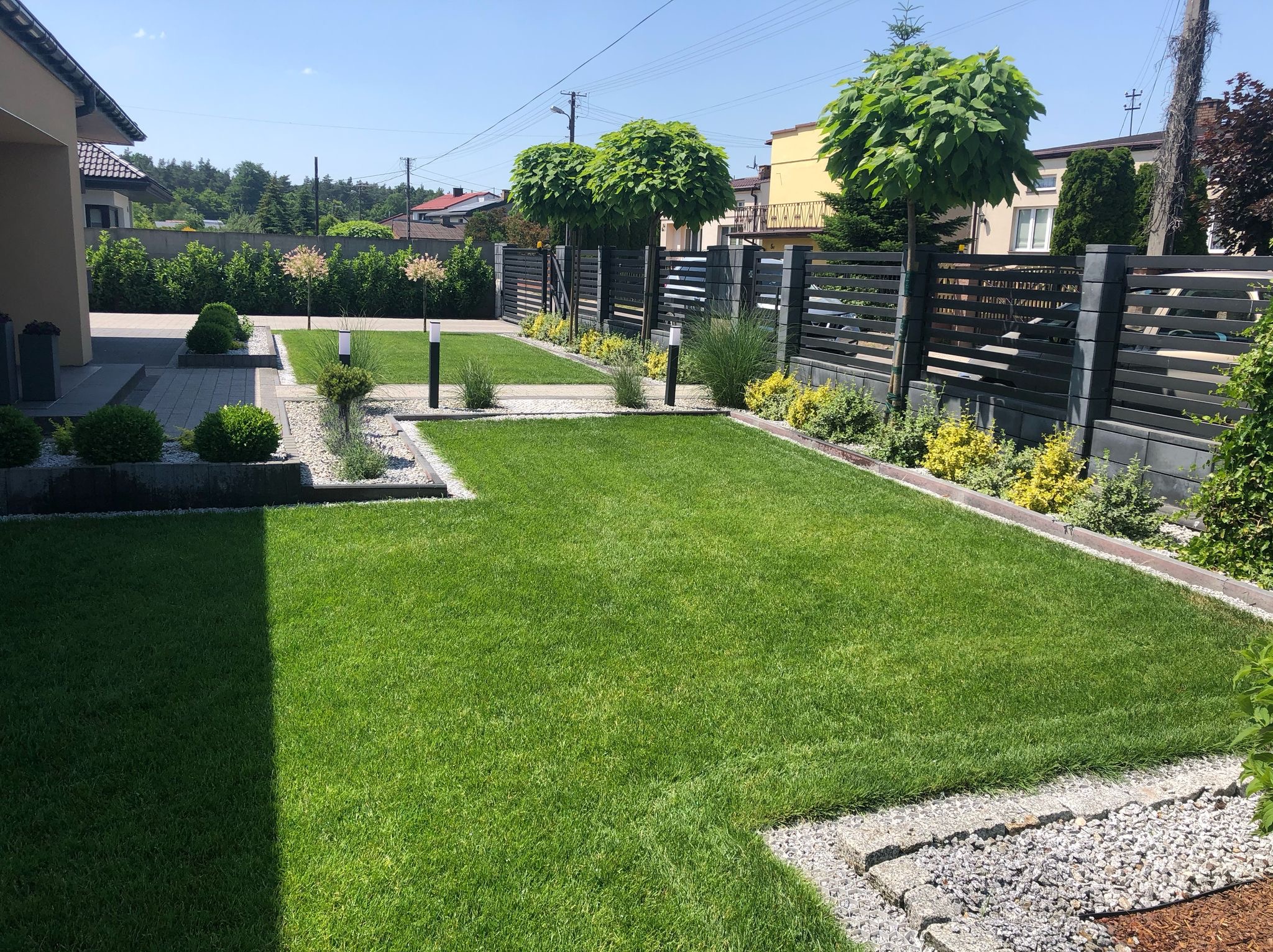 Zadbany trawnik z równo przystrzyżoną trawą, obrzeżami z kamienia i ozdobnymi drzewami w tle, oświetlony słońcem. Widoczne elementy architektury ogrodowej.