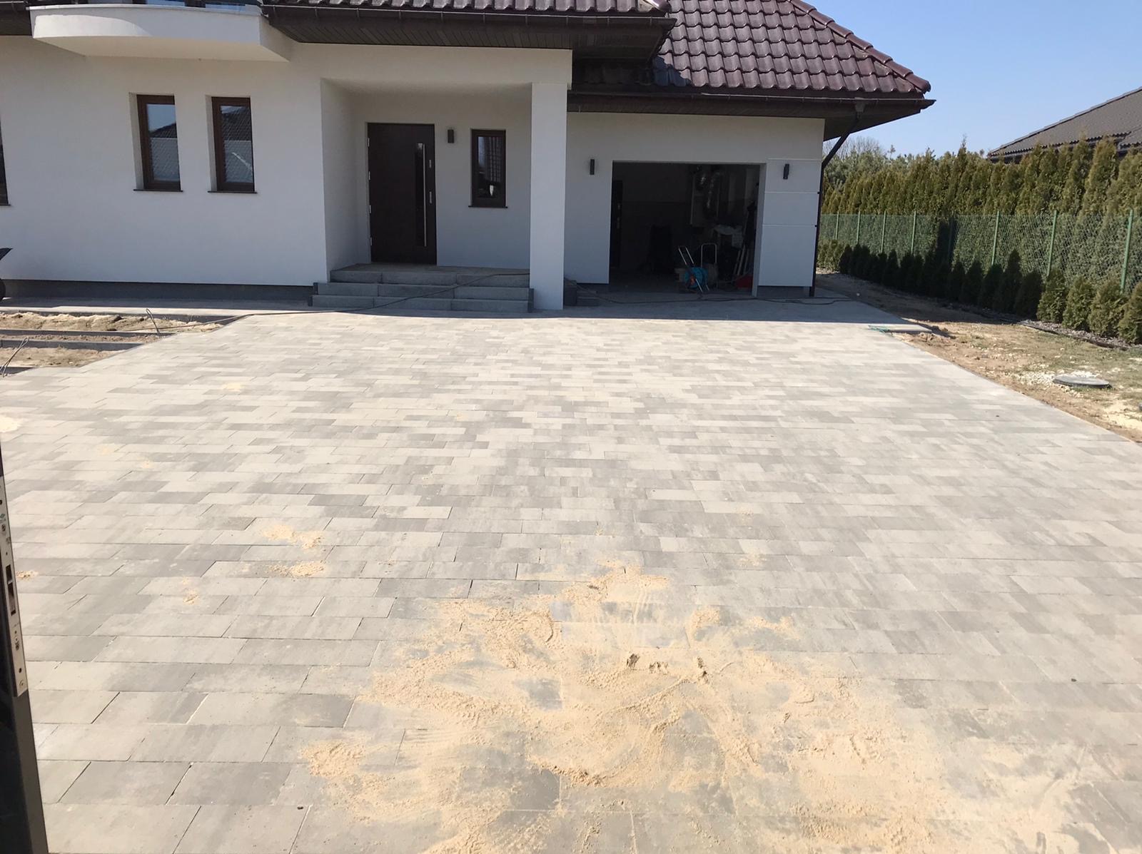 Nowa, szara kostka brukowa przed domem z białymi ścianami i brązowym dachem. W tle widoczne tuje i błękitne niebo. Częściowo zasypana piaskiem.