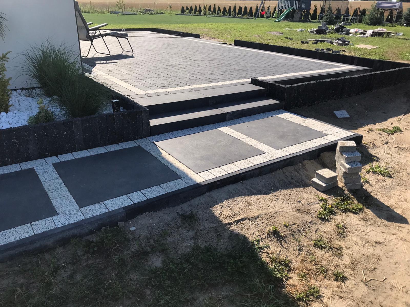 Nowoczesny taras z kostki brukowej w odcieniach szarości, z niskim murkiem i schodami. W tle widoczny trawnik i rząd drzew. Elementy dekoracyjne z białego kamienia.