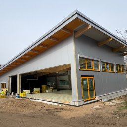 PHU Mateusz Cugier - Budowa hali z płyt warstwowych: szara elewacja, żółte ramy okien i drzwi, widoczne ocieplenie podłogi, pracownicy w odblaskowych kamizelkach. Hala w trakcie budowy.