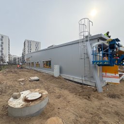 PHU Mateusz Cugier - Montaż hali z płyt warstwowych: ekipa na podnośniku, widoczne płyty, drabina na dachu, teren budowy z piaskiem i studzienkami. W tle bloki mieszkalne.