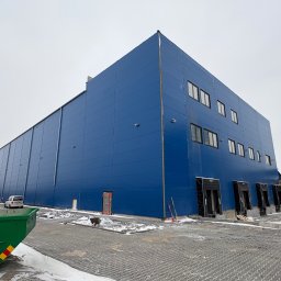 PHU Mateusz Cugier - Niebieska hala z płyt warstwowych w zimowej scenerii. Widoczne okna, bramy, kontener i sprzęt budowlany. Ujęcie z dołu podkreśla wielkość konstrukcji.