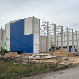 PHU Mateusz Cugier - Wznoszenie hali z betonu i płyt warstwowych, widoczny dźwig i materiały budowlane. Częściowo obłożona niebieską blachą trapezową. Etap budowy w toku.
