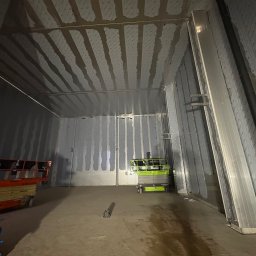 PHU Mateusz Cugier - Wnętrze hali w trakcie montażu płyt warstwowych. Widoczne podnośniki nożycowe i częściowo zamontowane płyty na ścianach i suficie. Surowy stan wykończenia.