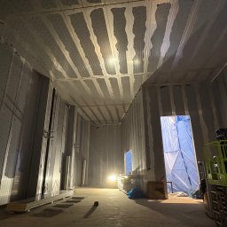 PHU Mateusz Cugier - Wnętrze hali w trakcie montażu płyt warstwowych na ścianach i suficie. Widoczny podnośnik nożycowy i materiały budowlane. Oświetlenie robocze.