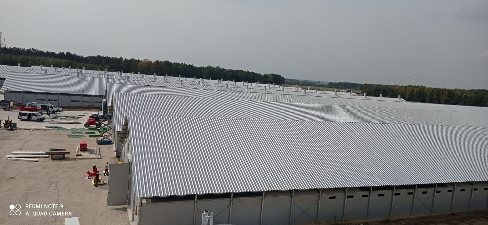 Hala z blachy trapezowej w trakcie budowy. Widoczne maszyny budowlane, elementy konstrukcyjne i pracownicy. W tle las i turbiny wiatrowe.