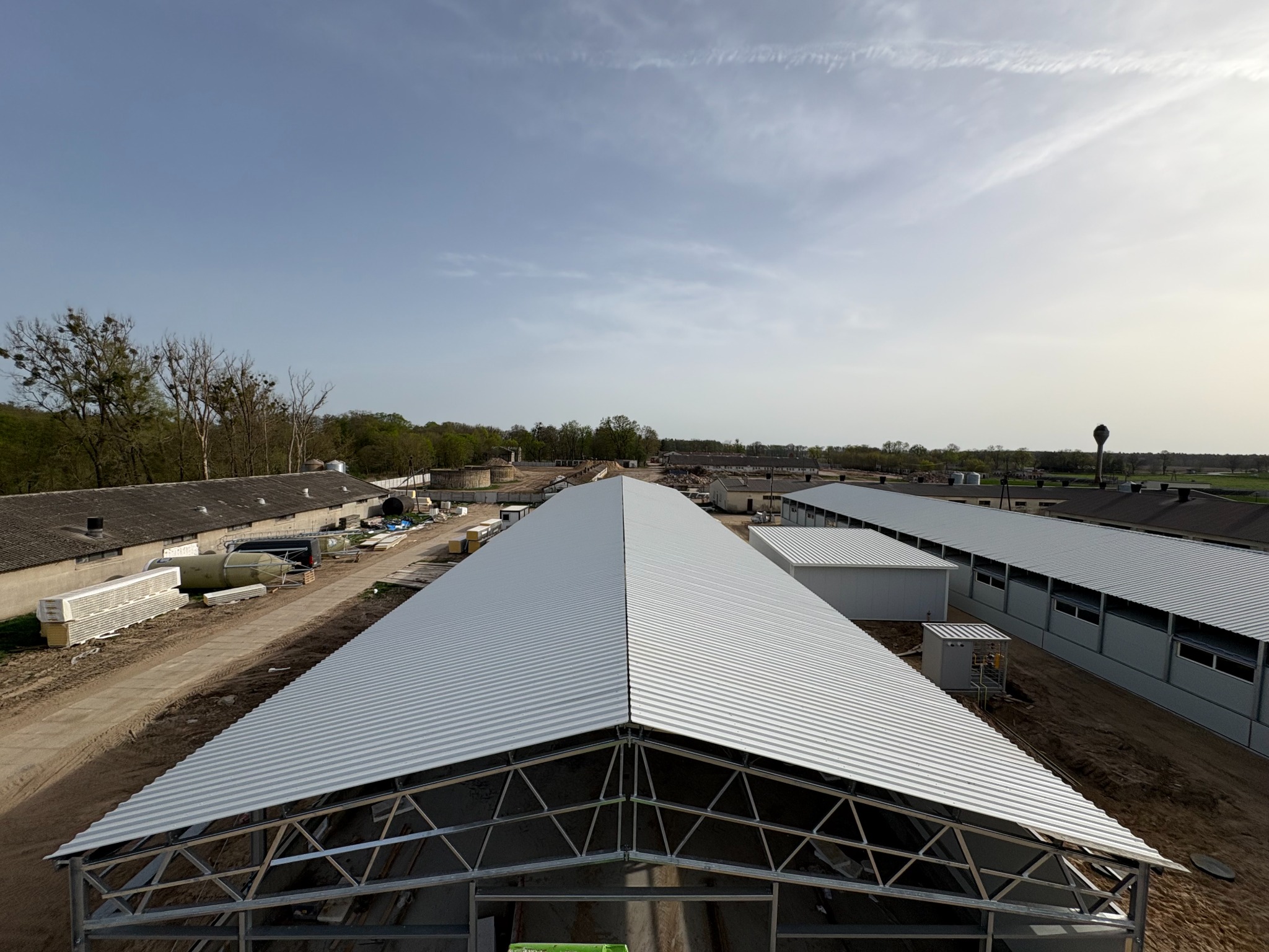 Wysoki kąt: hala w budowie z metalową konstrukcją dachu i białymi płytami warstwowymi w tle. Widoczne elementy krajobrazu i błękitne niebo.