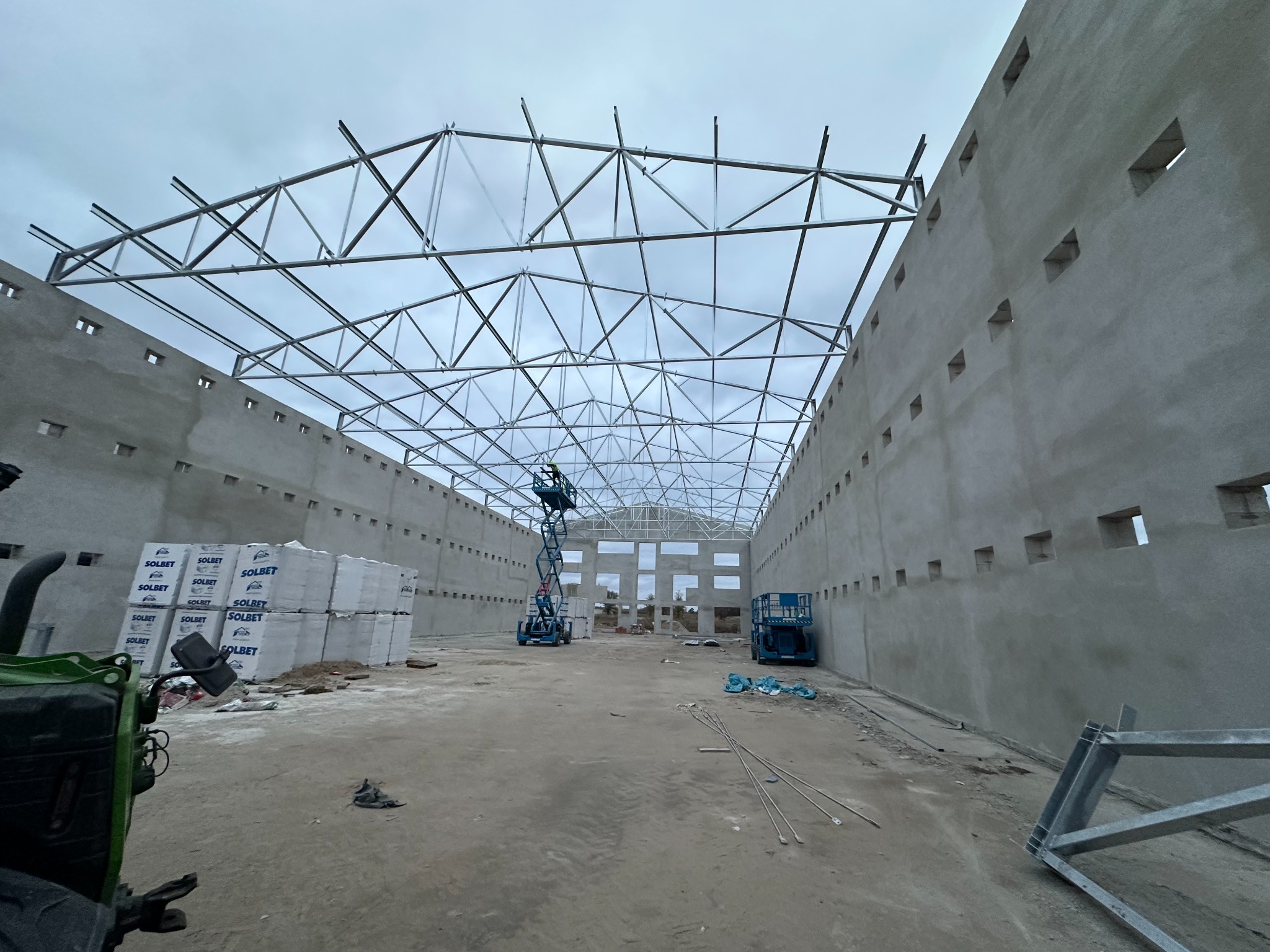 Budowa hali w toku: widoczne ściany z otworami, stalowa konstrukcja dachu, podnośniki i materiały budowlane. Ujęcie szerokokątne, pochmurne niebo.