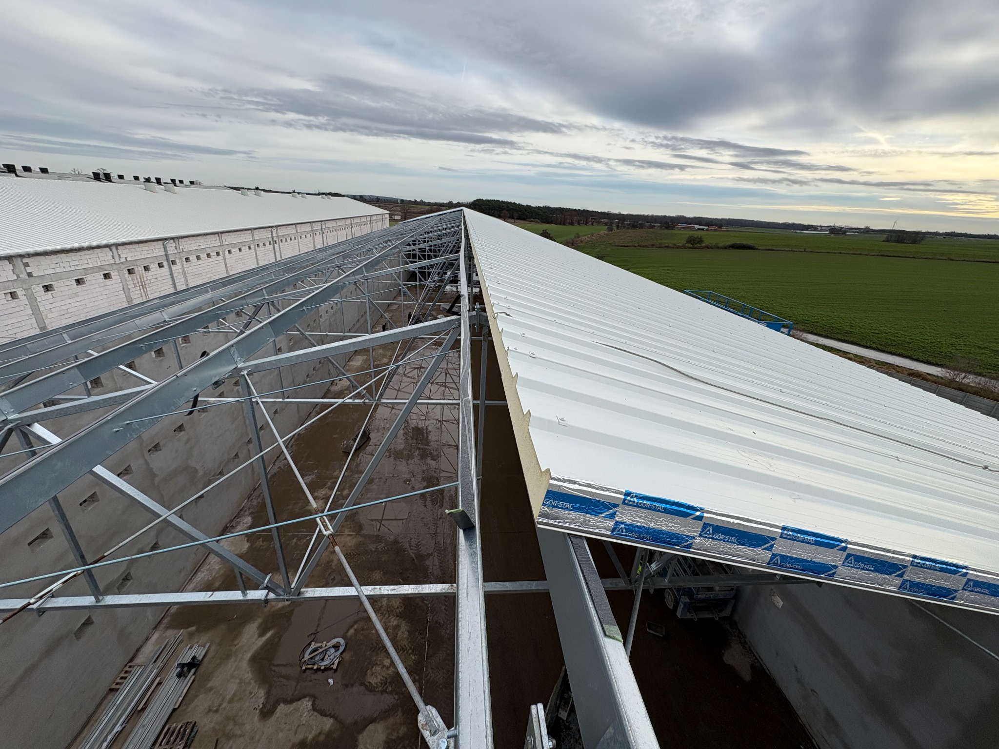 Budowa hali: montaż białych płyt warstwowych na stalowej konstrukcji dachu. Widok z góry na częściowo pokryty dach, pola w tle. Elementy mocujące widoczne.
