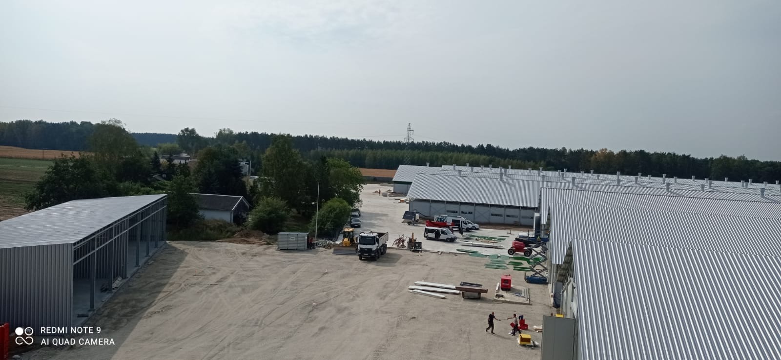 Widok z góry na teren budowy z halami o srebrnych dachach z blachy trapezowej, maszyny budowlane i dwóch pracowników na placu. W tle las i pola.