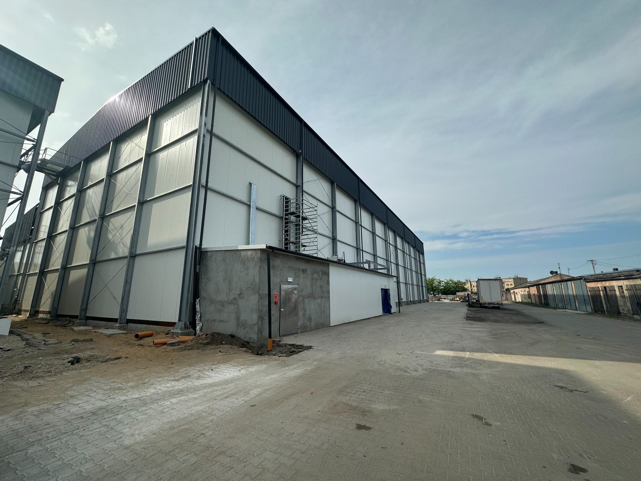 Nowoczesna hala z płyt warstwowych w trakcie budowy, widok z zewnątrz, metalowa konstrukcja, jasne ściany, fragment dachu, teren budowy z rurami.