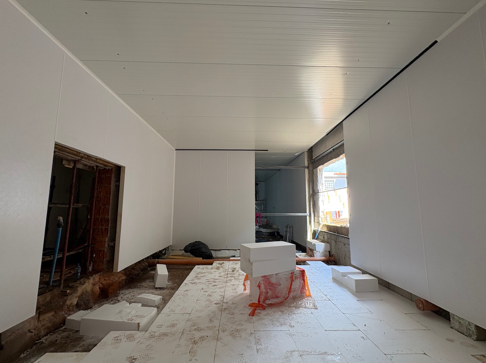 Wnętrze hali w trakcie montażu płyt warstwowych: białe płyty na ścianach i suficie, widoczne elementy konstrukcji i izolacji podłogi. Surowy stan wykończenia.