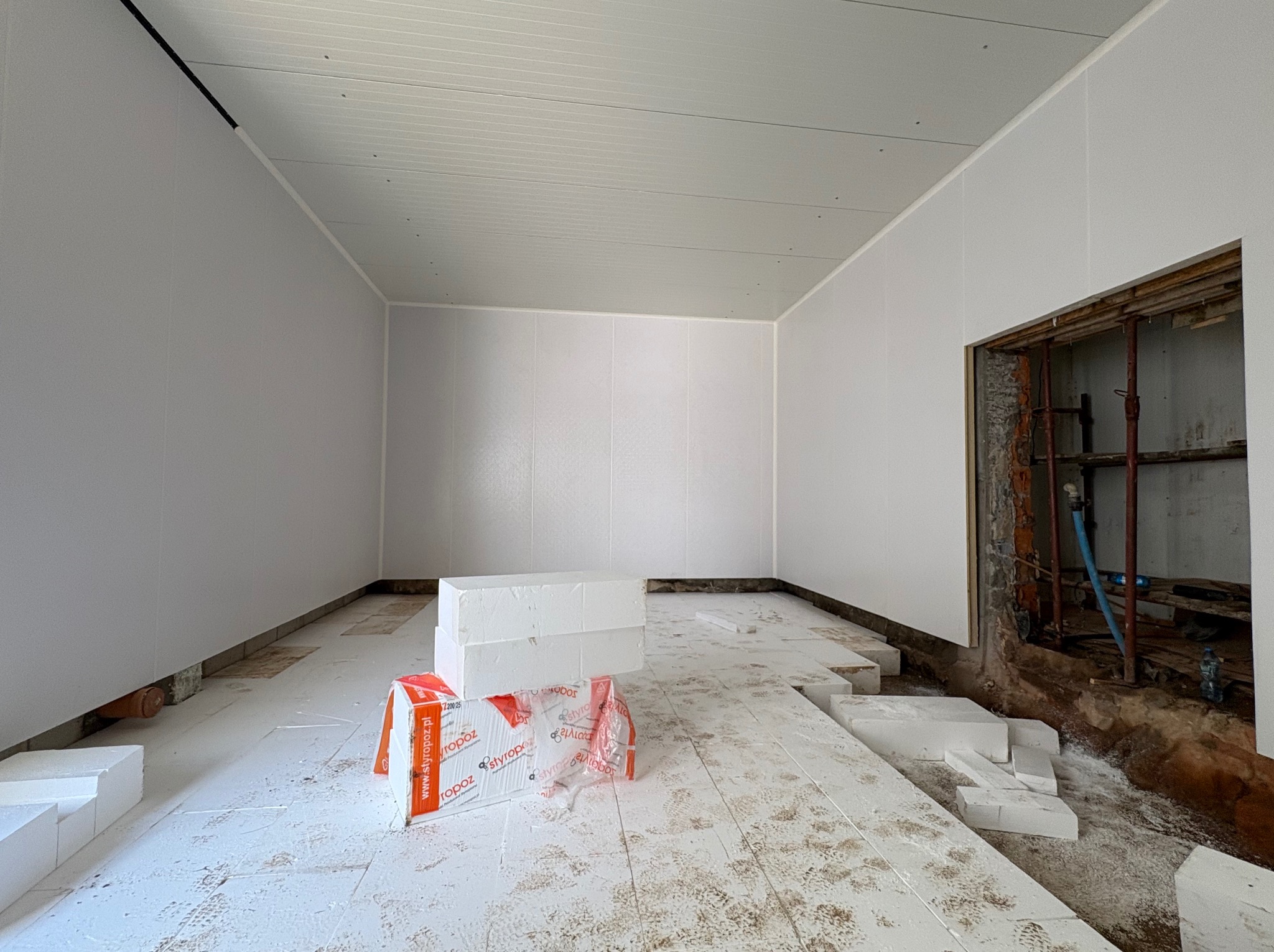 Wnętrze hali w trakcie montażu płyt warstwowych na ścianach i stropie. Na podłodze rozłożone płyty styropianowe i opakowania. Widoczne rusztowania w przejściu.