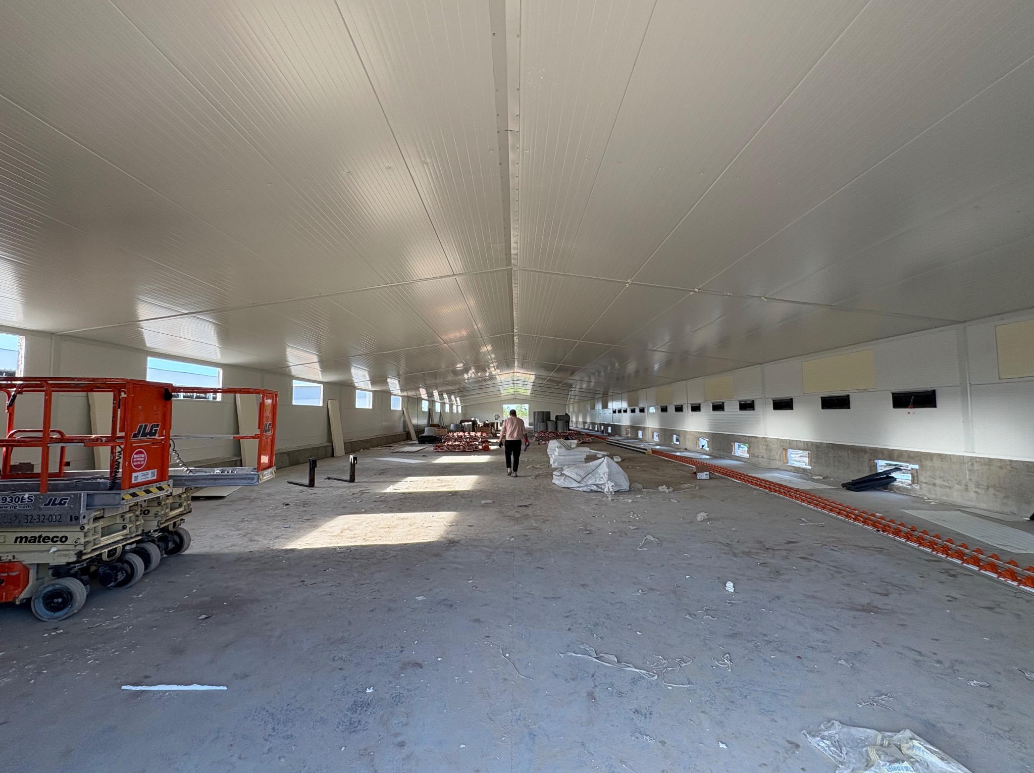 Wnętrze hali w trakcie budowy z zamontowanymi płytami warstwowymi na ścianach i suficie. Widoczny podnośnik i osoba w oddali.