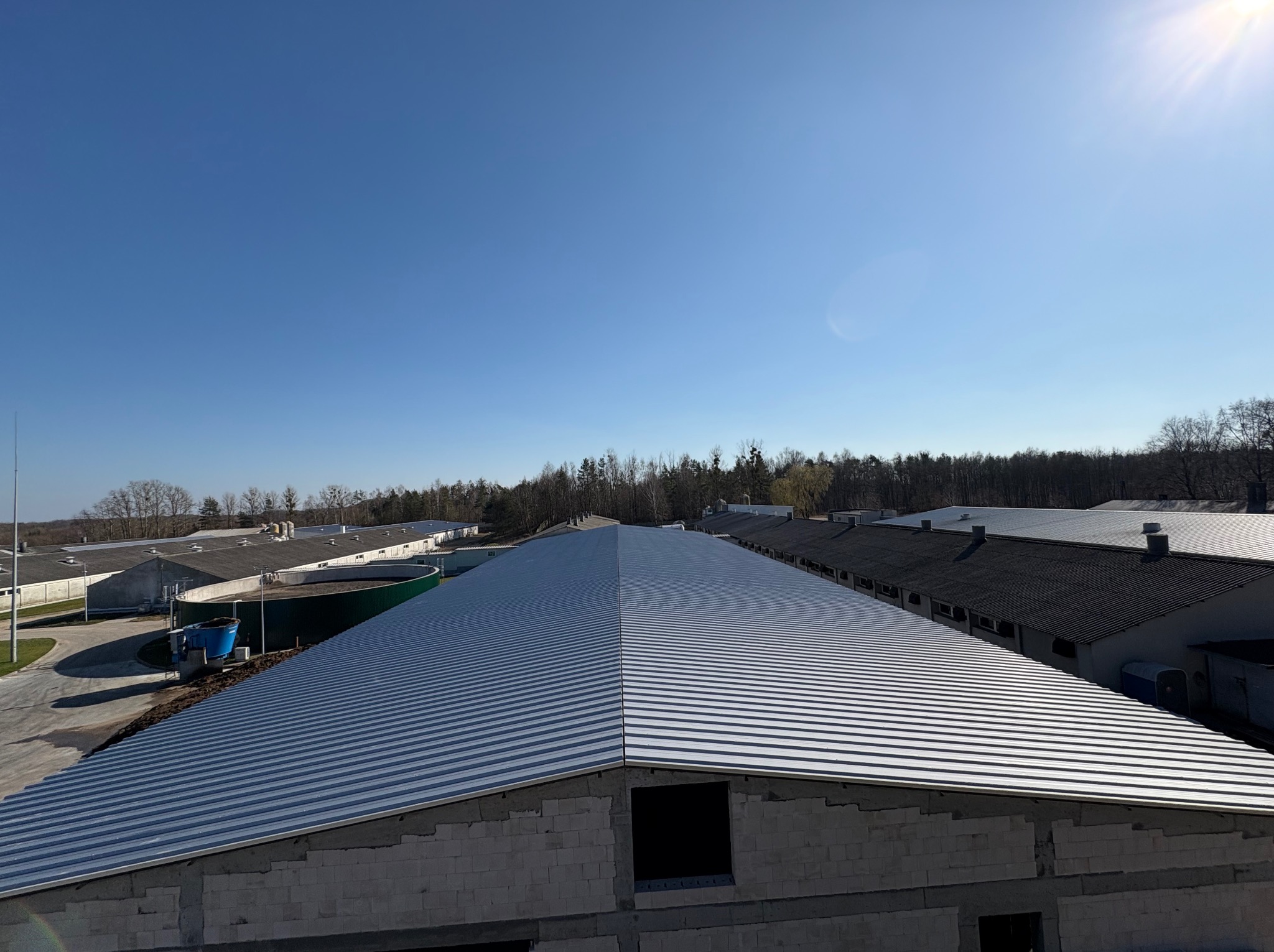 Hala w budowie z nowym, srebrnym dachem z blachy trapezowej. Widok z góry, w tle inne budynki i las. Słoneczny dzień.