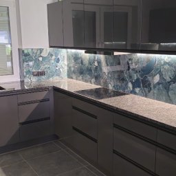 Marcin Sobusiak - Nowoczesna kuchnia z szarymi szafkami, blatem z konglomeratu i oknem PCV. Ściana nad blatem wykończona dekoracyjną płytą imitującą kamień.