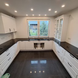 Marcin Sobusiak - Nowoczesna kuchnia w bieli z czarnym blatem i dużym oknem, widok na ogród. Ustawienie w kształcie litery U, ciemna podłoga. Elementy wykończenia w stylu minimalistycznym.