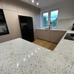 Kuchnia z beżowymi szafkami i blatem z konglomeratu w czarno-białe plamki. Widoczne okno, sprzęt AGD i drewniana podłoga.