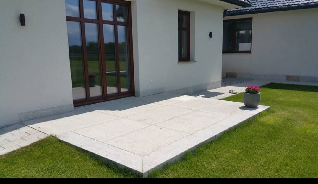 Taras z jasnych płyt, widok z poziomu trawnika. Brązowe drzwi przesuwne z szybami odbijają zieleń. Donica z kwiatami na wykończonym tarasie.